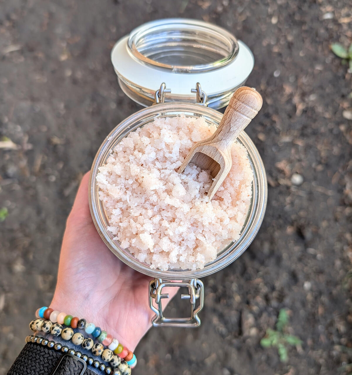 A hand holding a jar full of pink Gardener's Hand Scrub with a wooden scoop in it.
