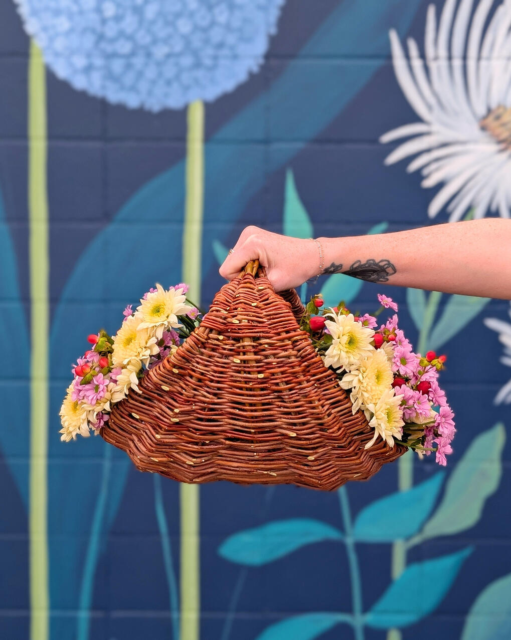 A hand holding a basket filled with flowers, against a blue wall.