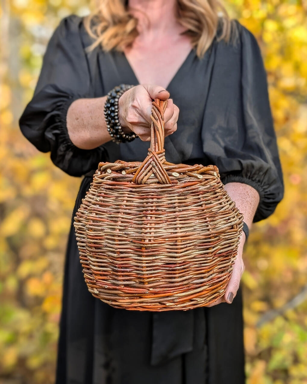 Women in black dress holding willow cauldron shaped basket with background of yellow leaves.